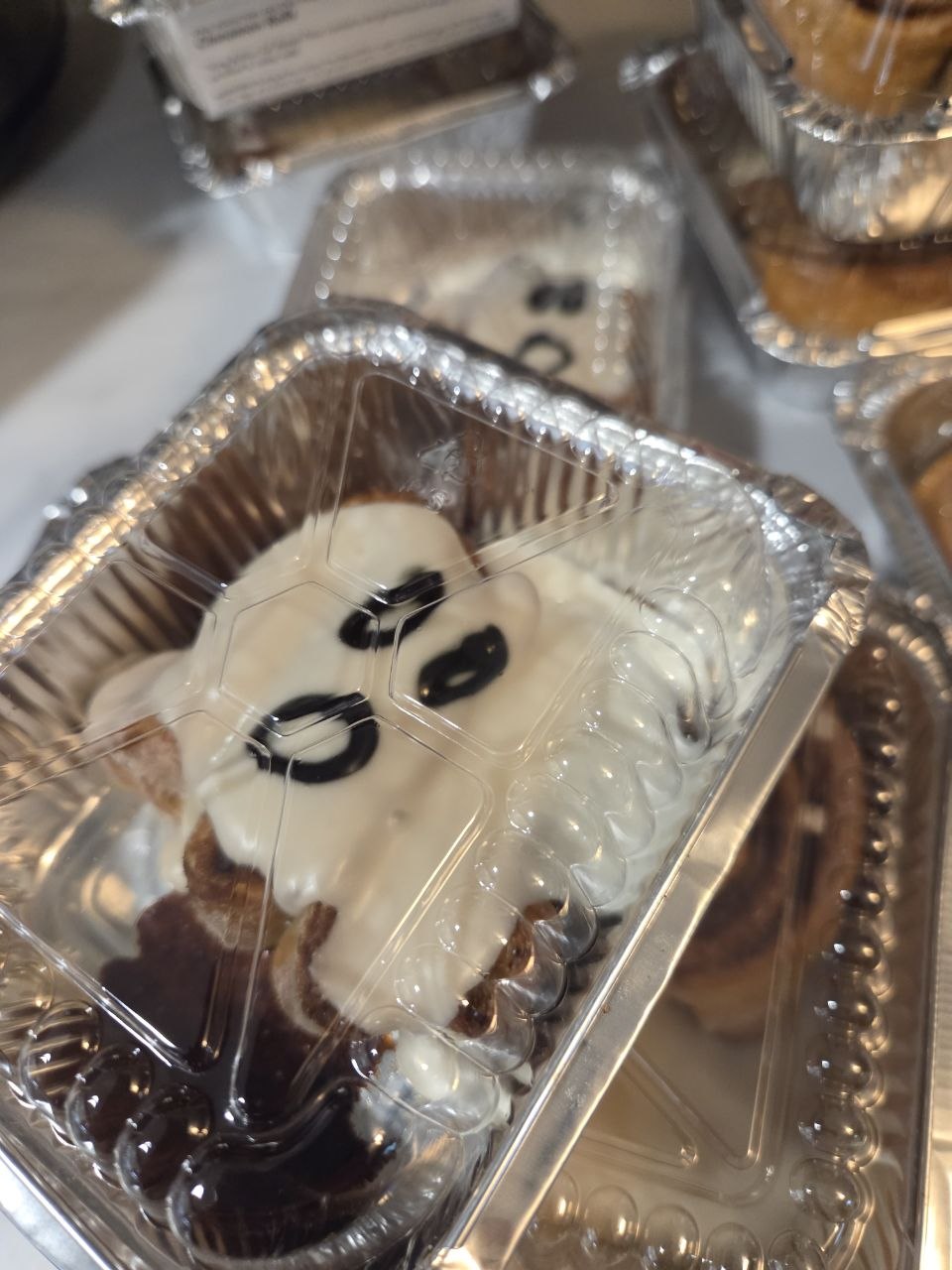 Cinnamon rolls with white glaze shaped like a ghost with black icing eyes, in a clear-lid foil pan
